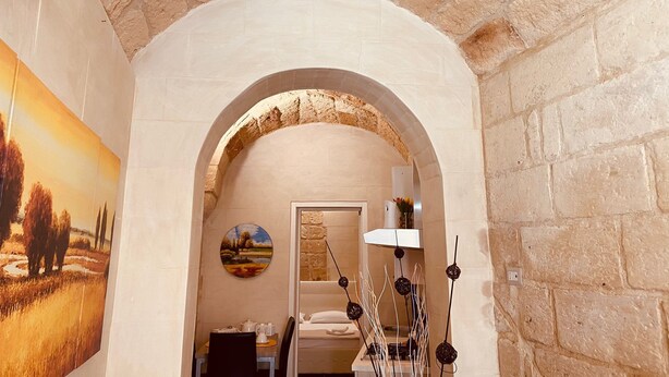 A narrow hallway with a stone wall and an arched doorway leading to a bedroom. A painting of a landscape is visible on the left wall, and a lamp is on the right.