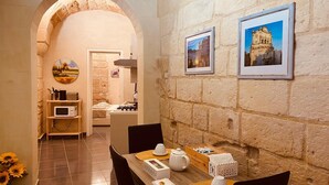 A cozy dining area with a stone wall, a wooden table set for two, and a view of a rustic kitchen in the background.