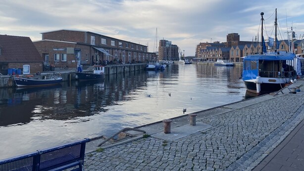 A canal with boats docked, buildings along the water, and a paved walkway. - Living in the historic imperial building (Wismar)