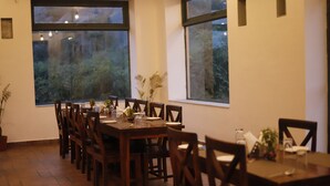 Un espace repas avec une longue table et des chaises en bois, de grandes fenêtres donnant sur un paysage verdoyant et une plante en pot posée sur le sol.