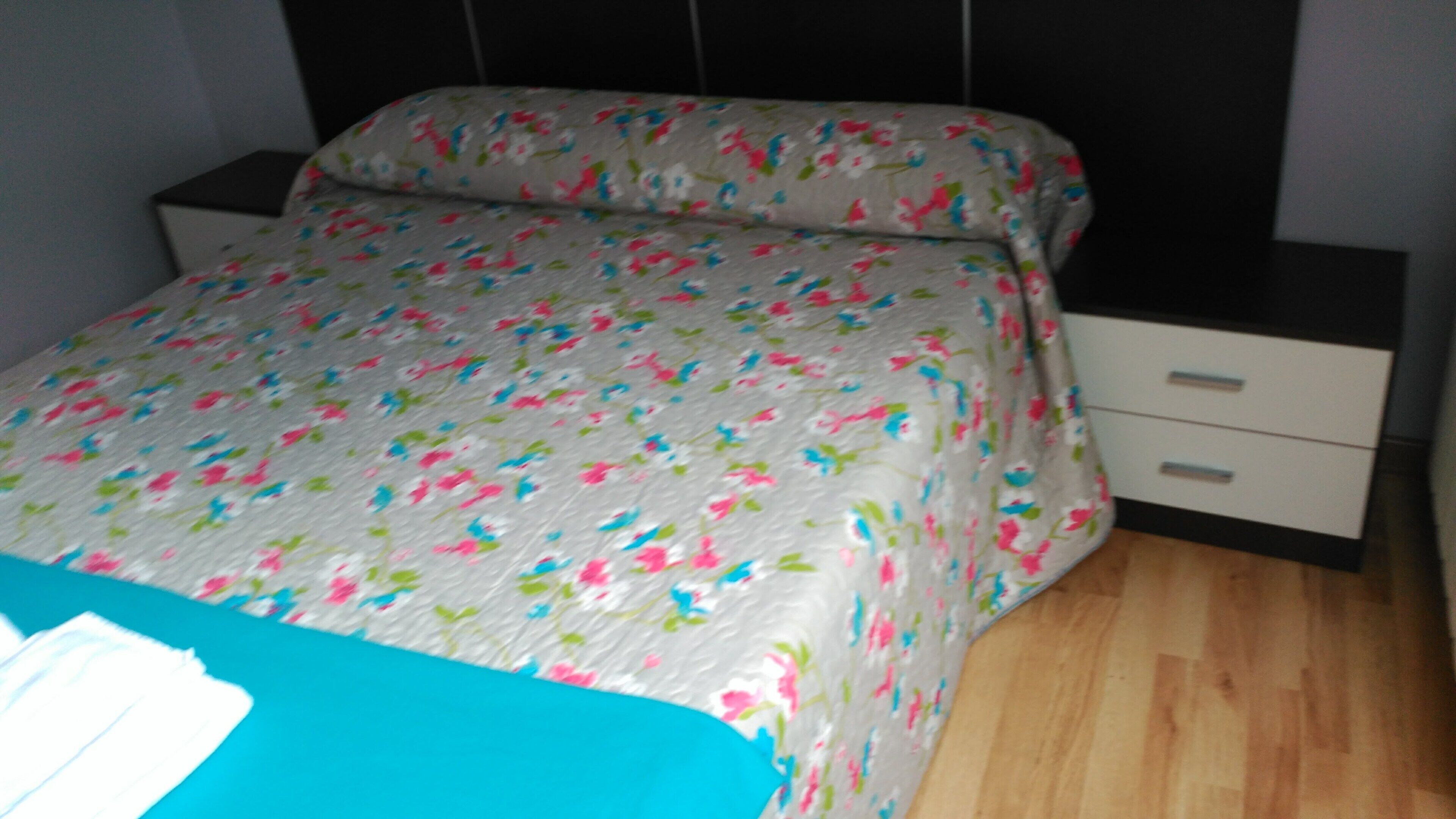 A bed with a floral bedspread, a nightstand with a drawer, and a wooden floor.