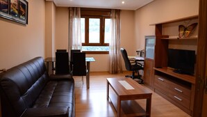 Uma sala de estar com sofá de couro preto, mesa de centro de madeira, televisão sobre suporte de madeira, escrivaninha com cadeira e janela com cortinas.