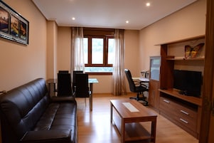 A living room with a black leather sofa, a wooden coffee table, a TV on a wooden stand, a desk with a chair, and a window with curtains.