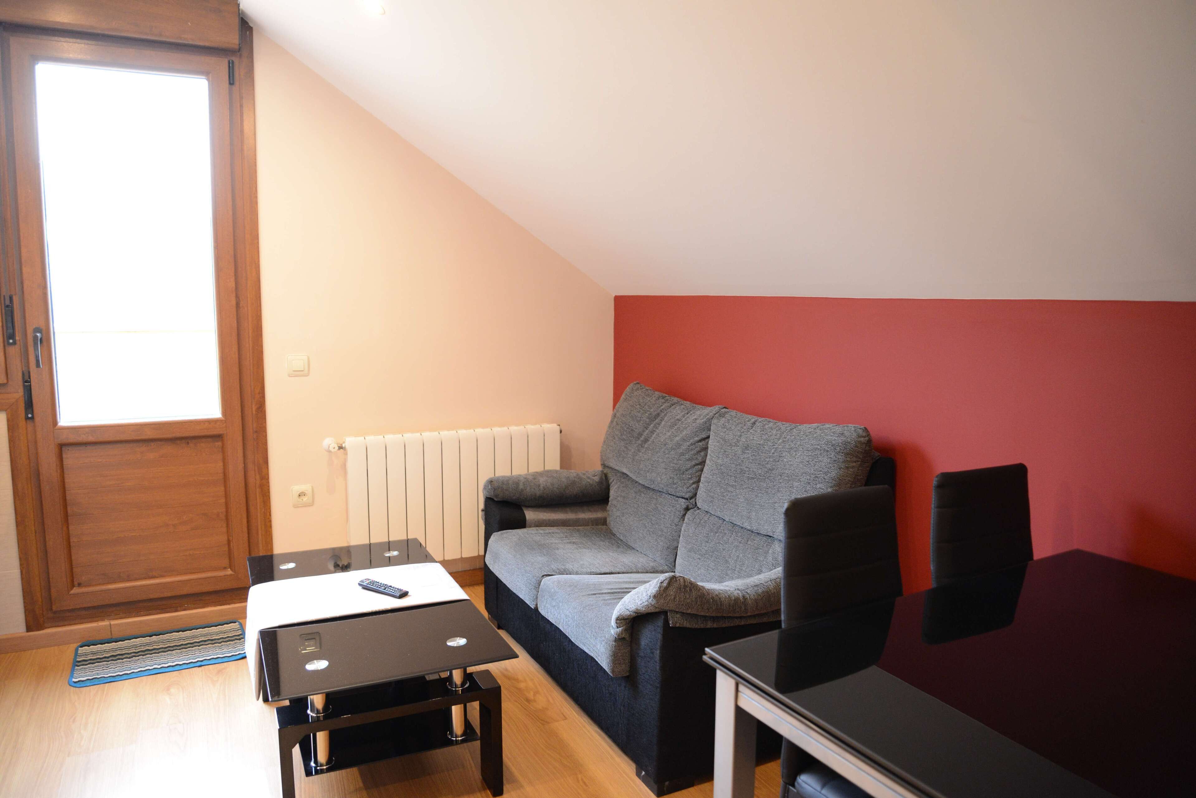 Una habitación pequeña y bien iluminada con una puerta de madera, un radiador, un sofá gris, una mesa negra y una pared roja.