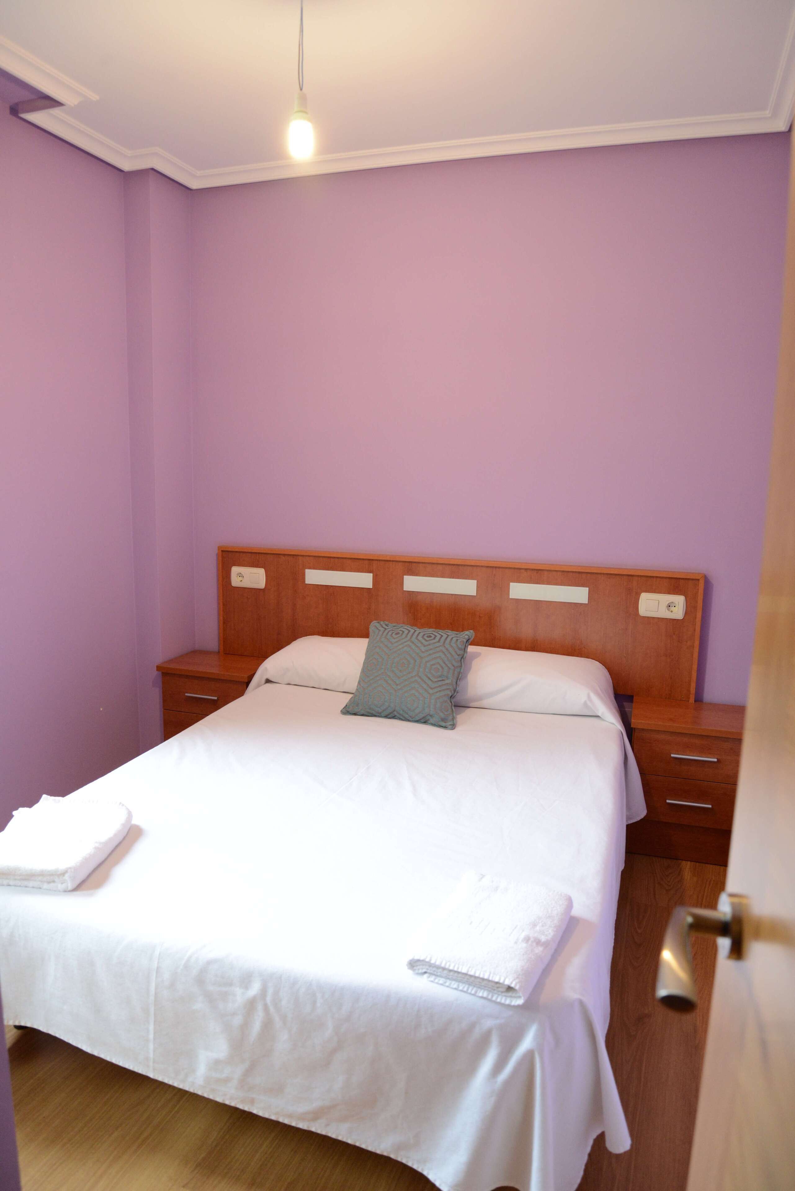 Una cama bien hecha con sábanas blancas y una sola almohada, una mesita de noche de madera con cajón, y una lámpara colgante arriba.