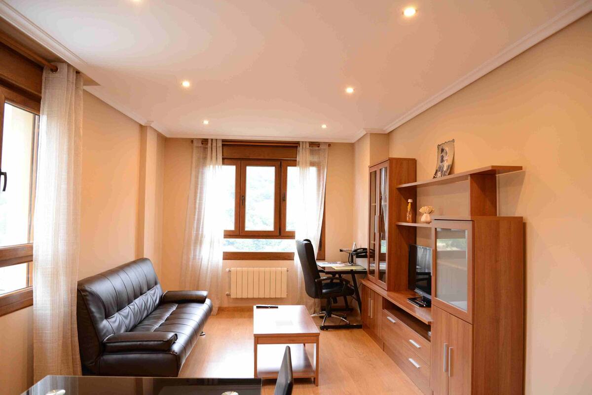 Uma sala de estar moderna com um sofá de couro, uma mesa de centro de madeira, uma escrivaninha com uma cadeira e uma televisão sobre um aparador de madeira.