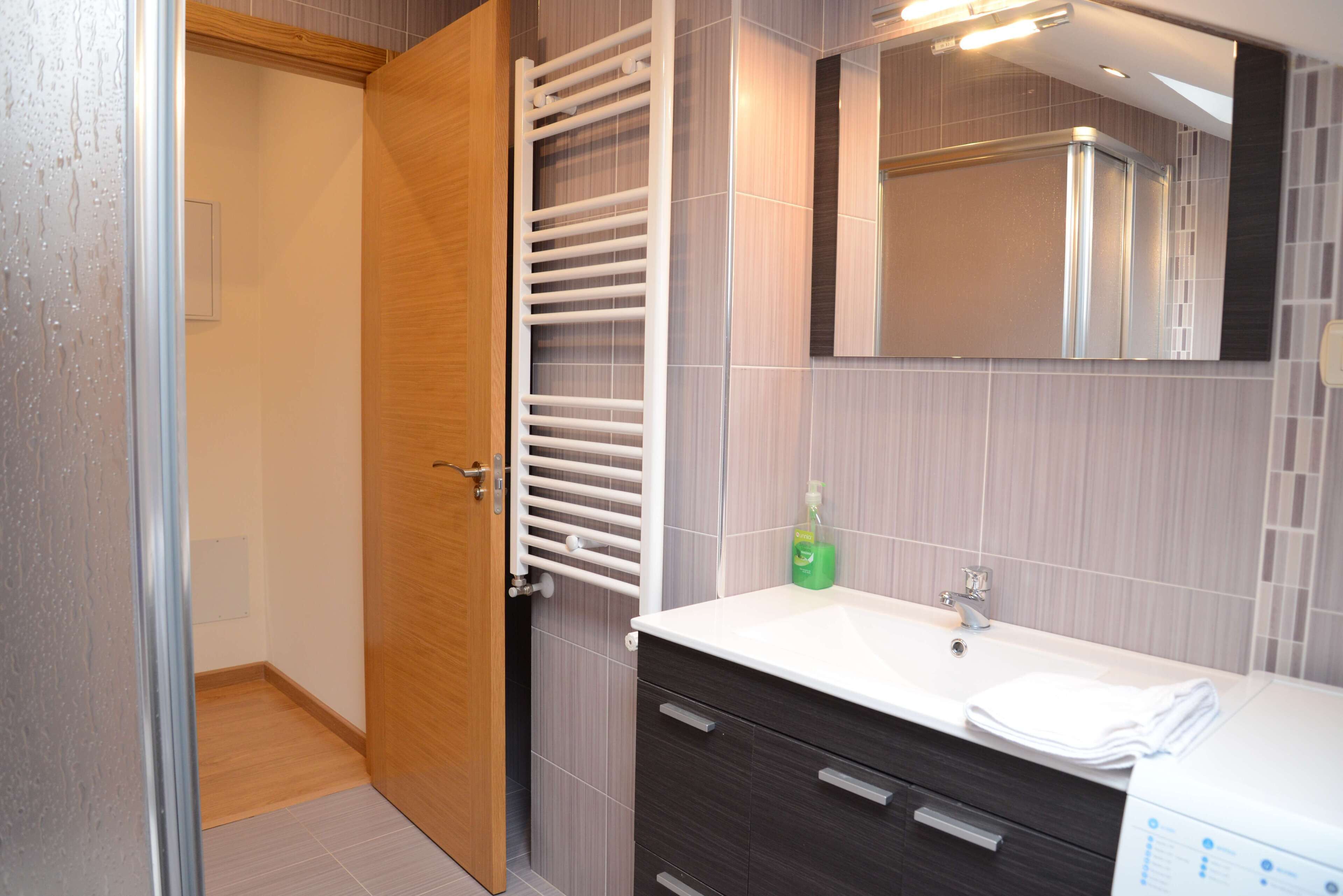 Un baño moderno con puerta de madera, toallero blanco, espejo y lavabo blanco con dispensador de jabón.