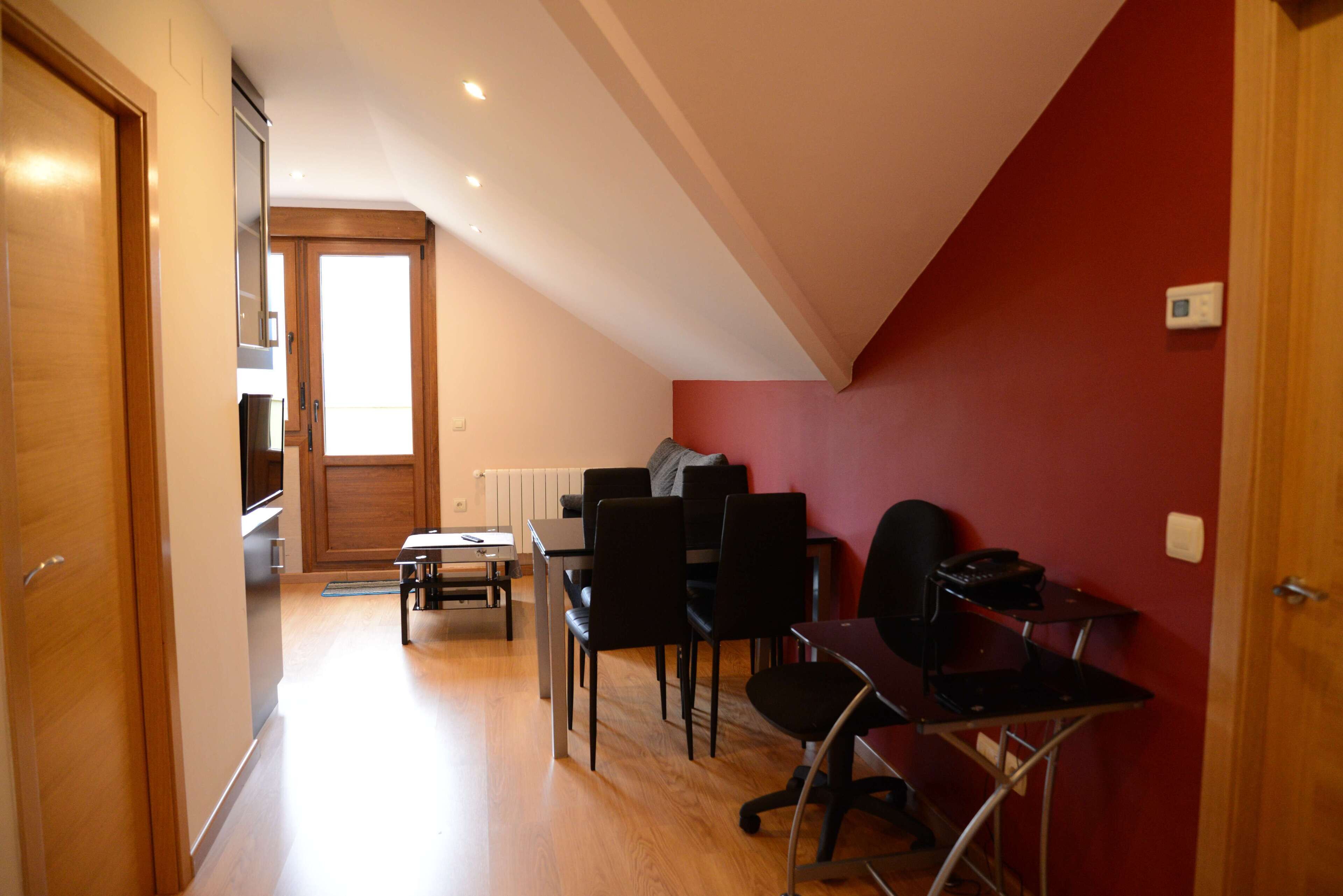 A room with a dining table, chairs, a desk, and a computer. There is a door on the left and a window at the end of the room.