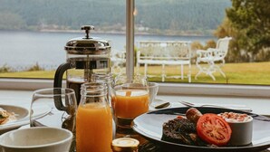 A breakfast table with a coffee maker, a glass of orange juice, and a plate of food, overlooking a lake and mountains. - Foyers Lodge (Inverness)