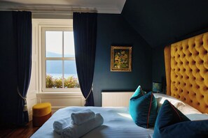 A bedroom with a large window, a bed with white linens, a yellow tufted headboard, and blue curtains. - Foyers Lodge (Inverness)