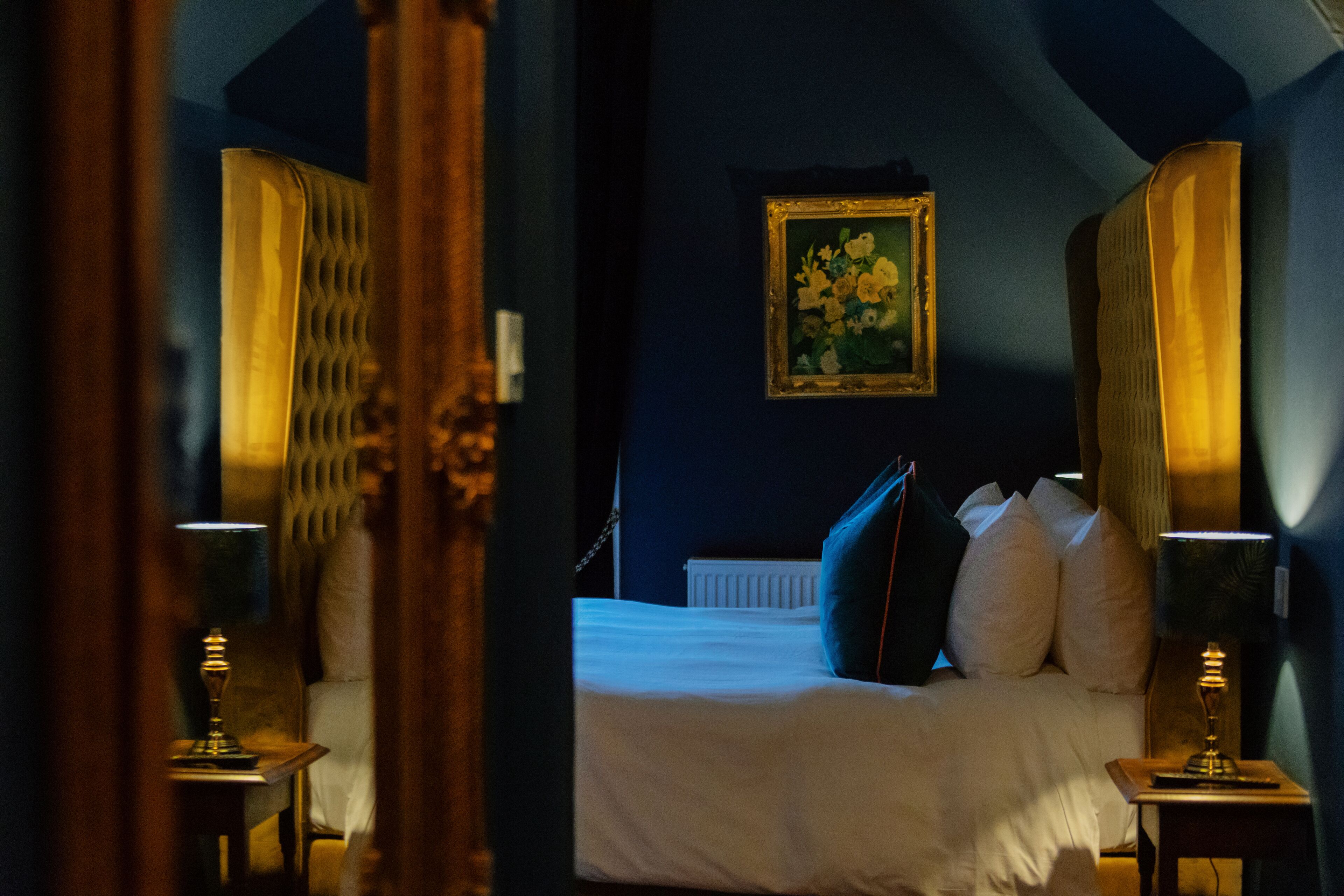 Un dormitorio con una cama, dos mesitas de noche, una mesita auxiliar, un cuadro enmarcado en la pared y una ventana con cortinas.