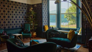 A room with patterned wallpaper, a large window with a view, and a green sofa with yellow cushions. - Foyers Lodge (Inverness)