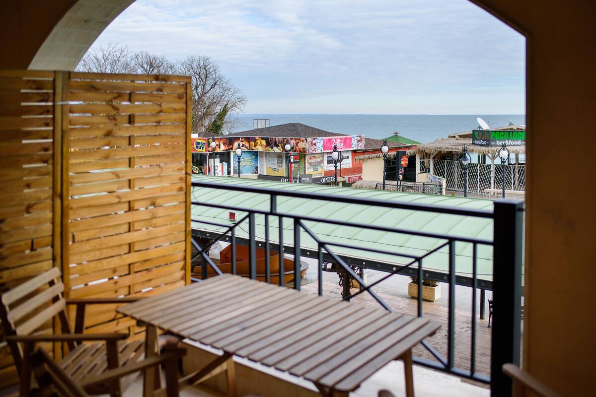 A balcony with wooden furniture, a view of the sea, and various colorful buildings in the background.