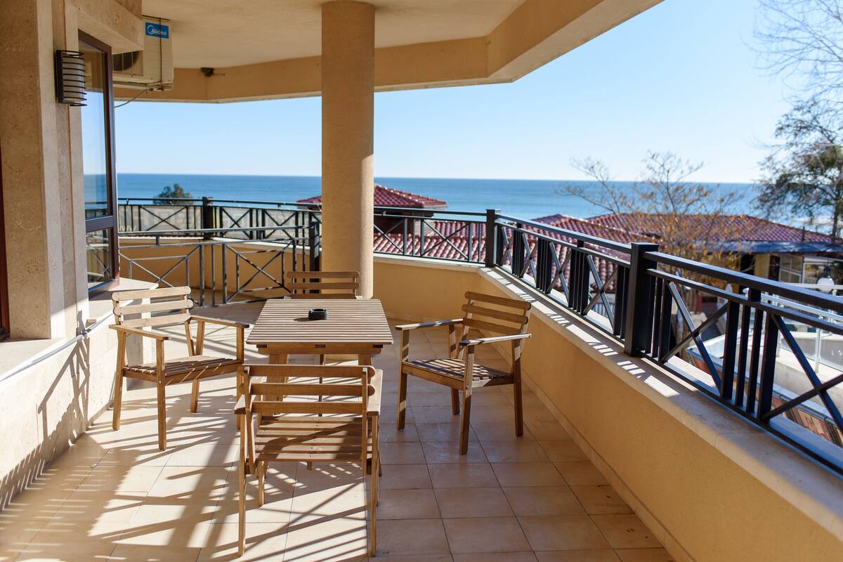 A balcony with wooden furniture, a view of the sea, and a building with red-tiled roofs.