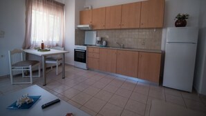 A compact kitchen with wooden cabinets, a white refrigerator, a microwave, and a dining table with chairs. - Dora Apartments (Malevizi)