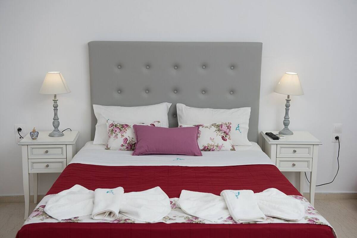 Cama bem arrumada com colcha vermelha, duas almofadas brancas e uma almofada decorativa roxa. Duas mesas de cabeceira com luminárias e um pequeno vaso em cada.