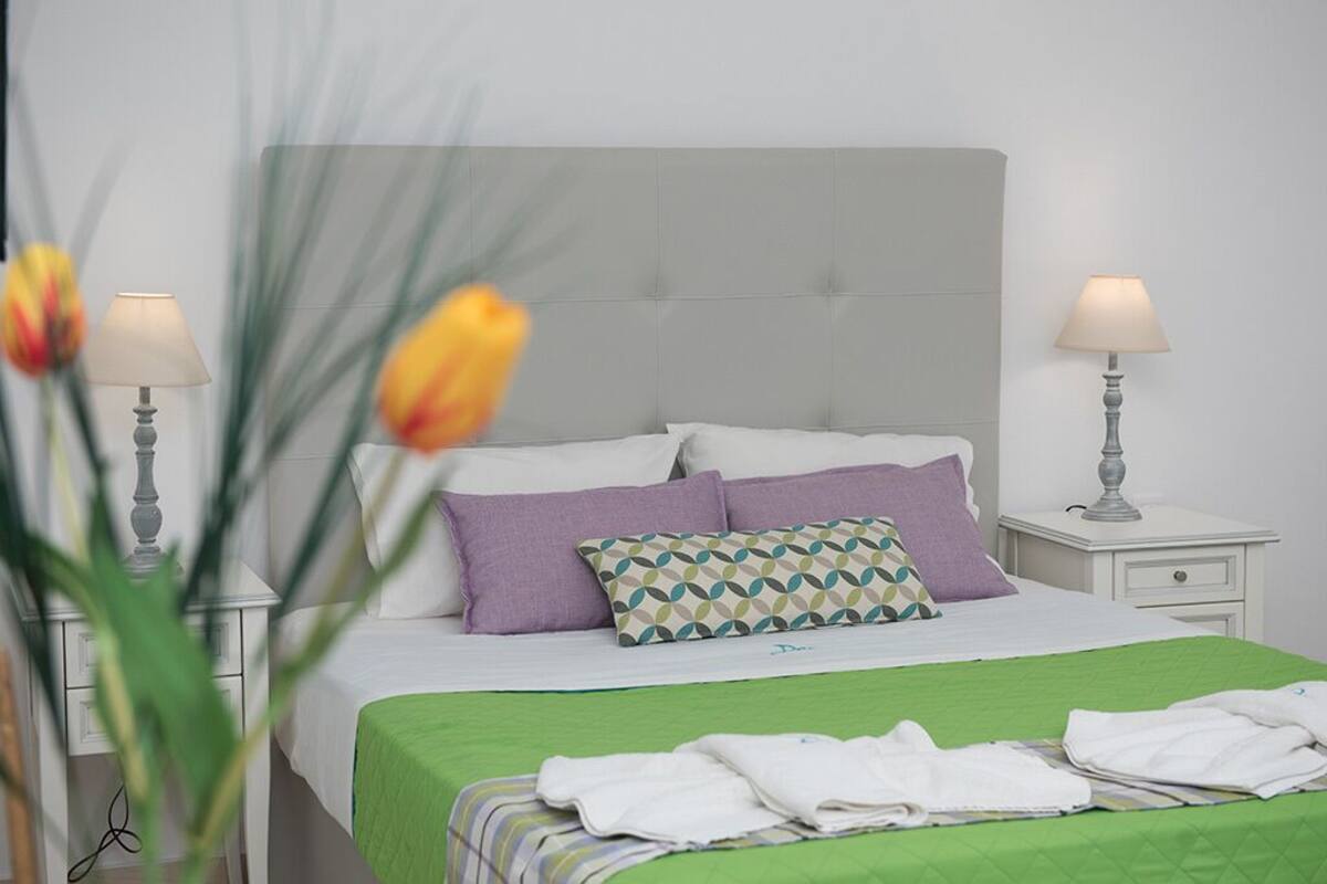 Cama bem arrumada com colcha verde, várias almofadas e mesa de cabeceira com abajur.