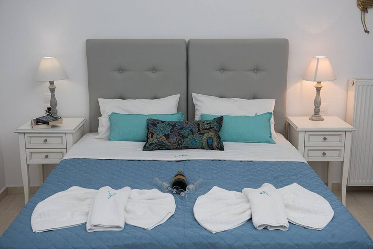 Uma cama de casal com cabeceira cinza, almofadas brancas e colcha azul. Há duas mesas de cabeceira com abajures e um livro pequeno em uma delas.