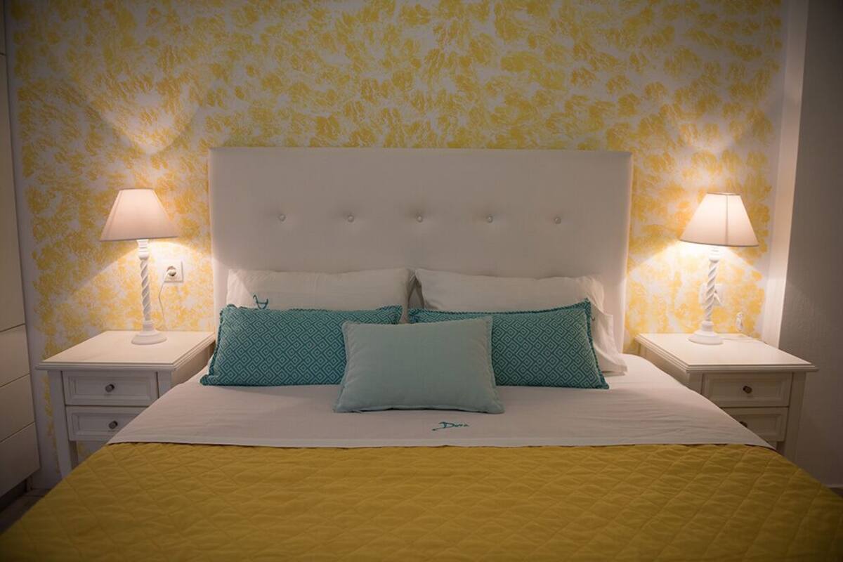 Cama bem arrumada com cabeceira branca e várias almofadas, duas mesas de cabeceira com abajures, e uma parede amarela com padrão.