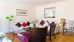 Ein modernes Wohnzimmer mit einem weißen Sofa, einem Glastisch und einem Sessel mit Zebra-Muster. An der Wand hängt ein gerahmtes Bild und über dem Sofa ein Spiegel.