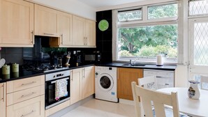 A modern kitchen with wooden cabinets, a built-in oven, a microwave, a washing machine, and a window with a view of greenery. - 4-bedroom Villa on Marble Arch (London)