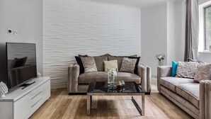 Een moderne woonkamer met een beige bank, een glazen salontafel en een flatscreen televisie op een witte kast.