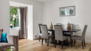 Een eetgedeelte met een tafel en stoelen, een raam met gordijnen, en een lijst met een afbeelding aan de muur.