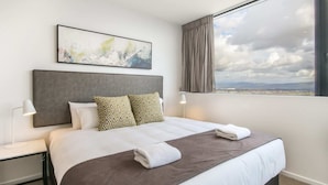 A hotel room with a large bed, a headboard, a window with a view, a painting on the wall, and a nightstand with a lamp. - Luxurious 2 Bedroom Brand New Apartment With Amazing Hinterland Views (Broadbeach)