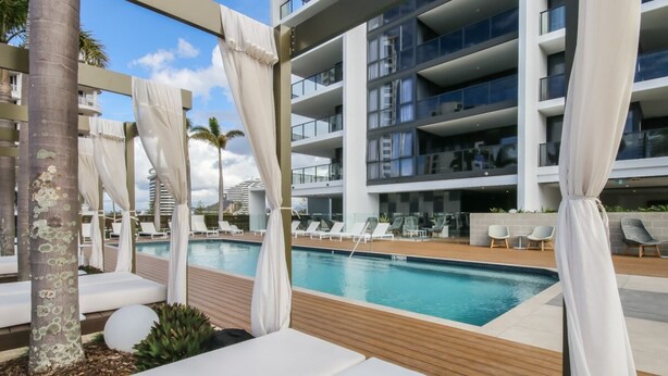 A modern hotel pool area with white lounge chairs, a wooden deck, and a view of the building's balconies. - Luxurious 2 Bedroom Brand New Apartment With Amazing Hinterland Views (Broadbeach)