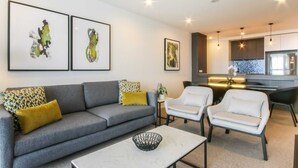 A modern living room with a grey sofa, white armchairs, and a marble coffee table. There are abstract paintings on the wall and a kitchen area in the background. - Luxurious 2 Bedroom Brand New Apartment With Amazing Hinterland Views (Broadbeach)