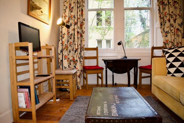 A cozy living room with a wooden bookshelf, a red cushioned chair, a small table, and a sofa with a checkered pillow.