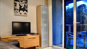 A modern living room with a flat-screen TV on a wooden cabinet, a glass coffee table, and a sliding glass door leading to an outdoor area with a chair and table. - Minimal Studio With Views Over Battersea Park (London)