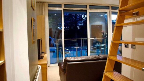 A modern hotel room with a leather sofa, a wooden ladder, and a view of an outdoor pool area. - Minimal Studio With Views Over Battersea Park (London)