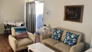 Ein Wohnzimmer mit beiger Couch, zwei Sesseln, einem kleinen weißen Tisch und einem Bild an der Wand.