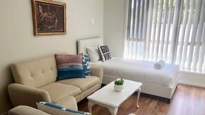 Ein Wohnzimmer mit einem beigen Sofa, einem weißen Sessel, einem weißen Couchtisch mit einer Pflanze und einem Bett mit weißer Bettwäsche und einem Kissen mit dem Aufdruck 'GOOD VIBE'.