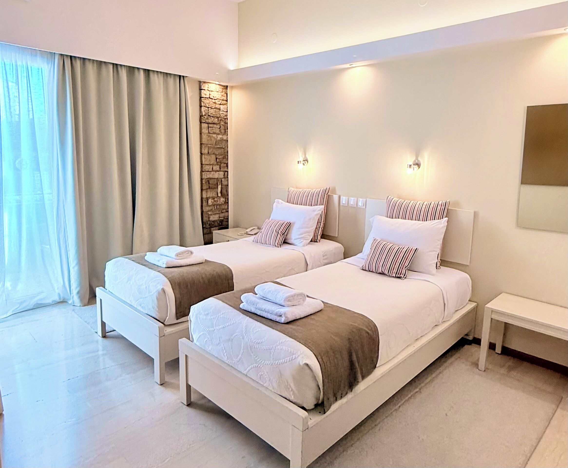 A hotel room with two beds, a stone accent wall, and a large window with curtains.