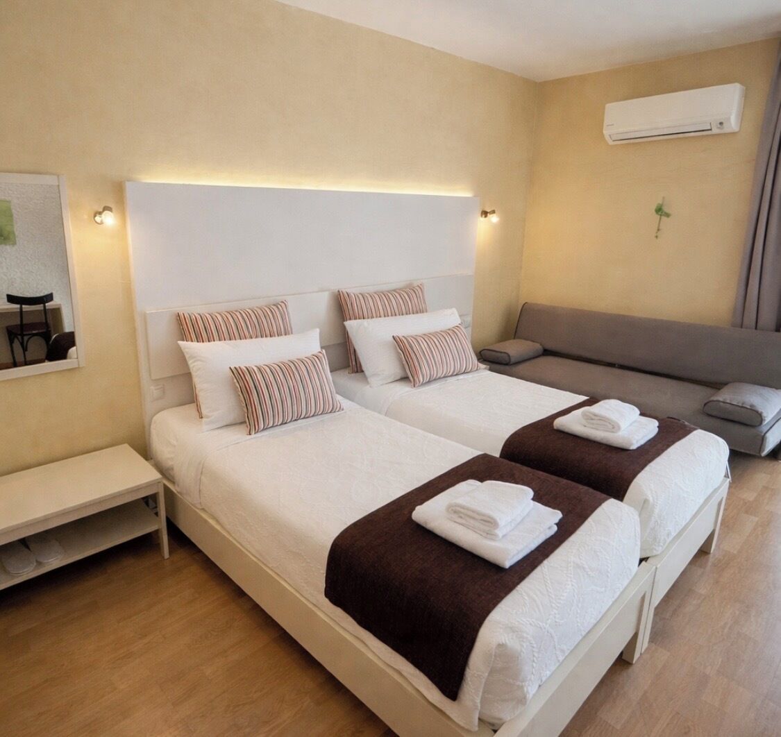 A hotel room with two beds, a sofa, a bench, and an air conditioning unit on the wall.