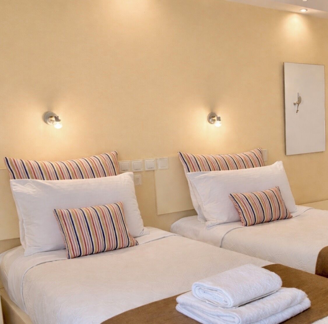 Habitación de hotel con dos camas, cada una con almohadas a rayas y ropa de cama blanca. Sobre cada cama hay una lámpara de pared y un cuadro en la pared.