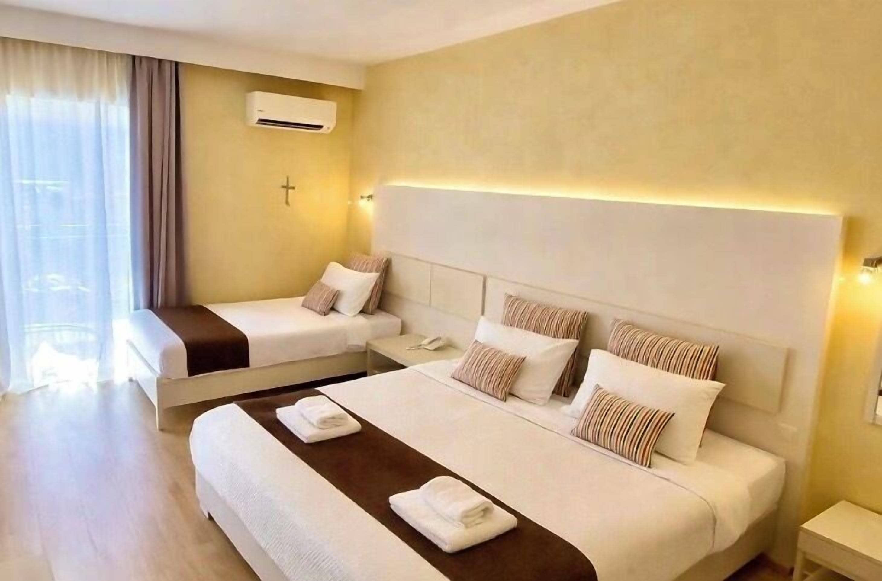 A hotel room with two beds, a large window with curtains, an air conditioning unit, and a bedside table with a phone.
