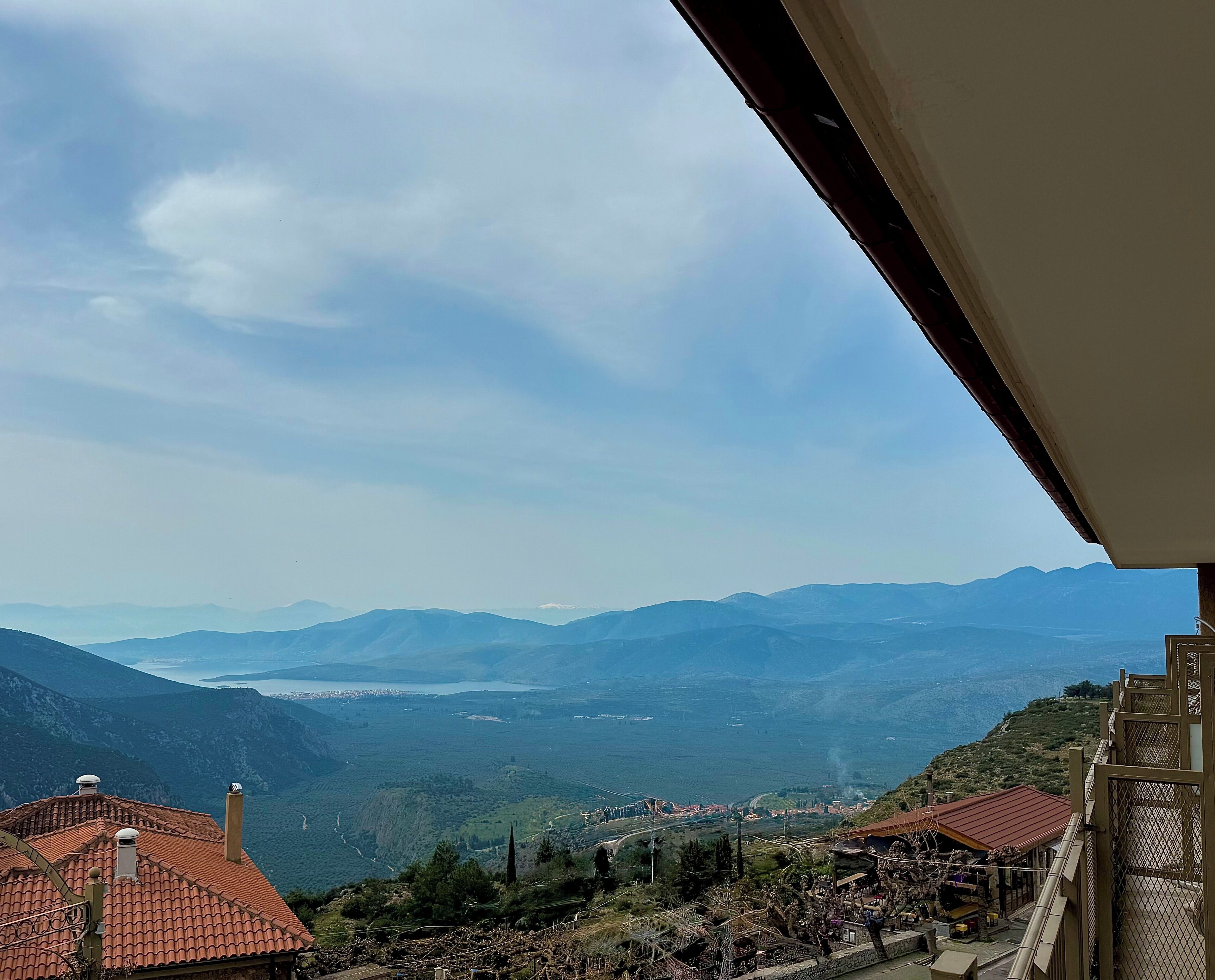 Vista desde un balcón sobre un valle con montañas a lo lejos, una masa de agua y un pueblo pequeño abajo.
