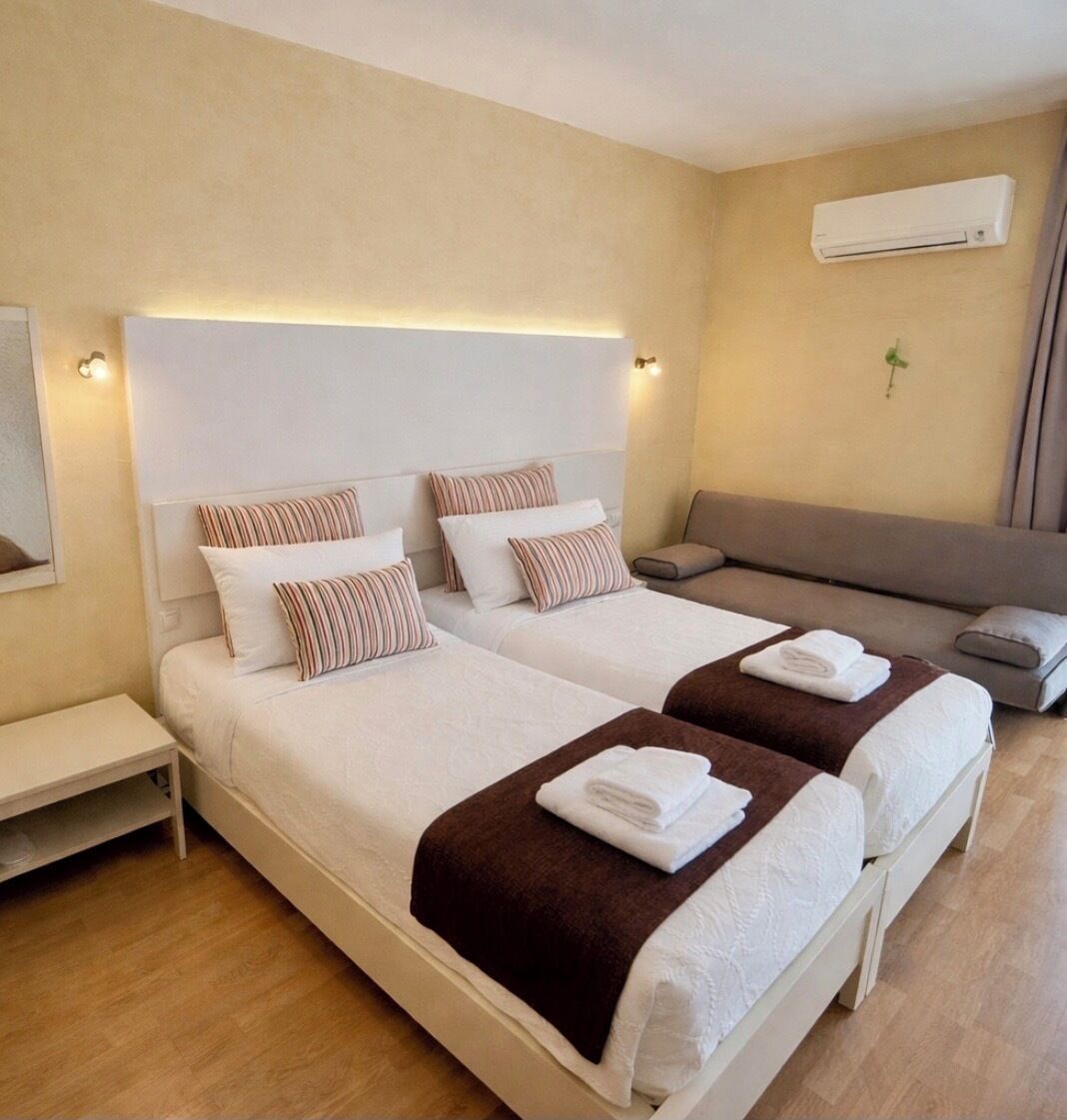 Quarto de hotel com duas camas, um sofá, um aparelho de ar condicionado e uma mesa de cabeceira.