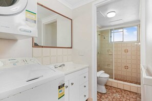 Baño con lavadora, lavamanos, espejo, inodoro y zona de ducha.