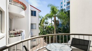 A balcony with a round table and chairs, a small garden area with palm trees, and a view of neighboring buildings. - Superb 1 Bedroom Apartment Near the Beach (Burleigh Heads)