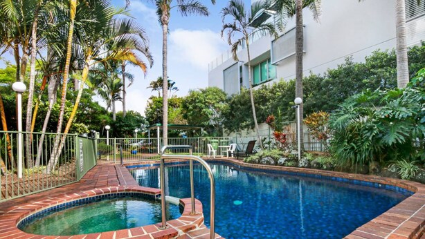 A swimming pool with a brick border, surrounded by palm trees and a white building. - Superb 1 Bedroom Apartment Near the Beach (Burleigh Heads)