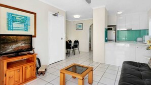A living room with a TV, wooden cabinet, and a small table. There is a dining area with chairs and a kitchen with cabinets and a microwave. - Superb 1 Bedroom Apartment Near the Beach (Burleigh Heads)