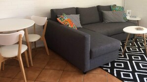 Een moderne woonkamer met een grijze bank, witte stoelen, een klein rond tafeltje en een kamerkleed met een patroon.