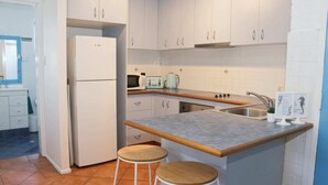Een compacte keuken met witte kasten, een inbouwoven, een magnetron, een waterkoker en een spoelbak. Er staan twee houten krukken bij het aanrecht.