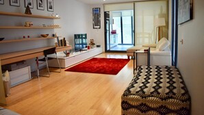Un salon moderne avec un bureau en bois, un tapis rouge, un canapé à motifs et une grande fenêtre donnant sur l’extérieur.