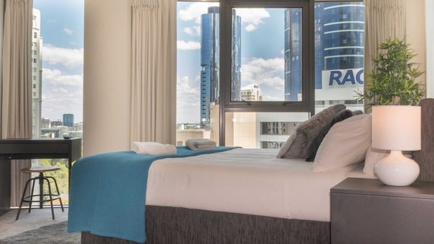 A hotel room with a large bed, a desk, and a view of tall buildings. - 1 Bedroom Studio Apartment near Eagle Street (Brisbane)