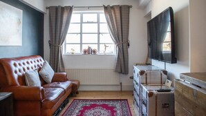 Ett mysigt vardagsrum med en lädersoffa, ett stort fönster med gardiner, en platt-TV och gamla resväskor.